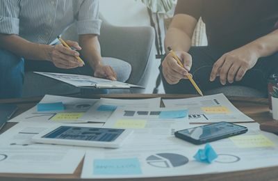 Zwei Männer sitzen zusammen am Tisch, vor ihnen viele Dokumente und erstellen ein Messekonzept